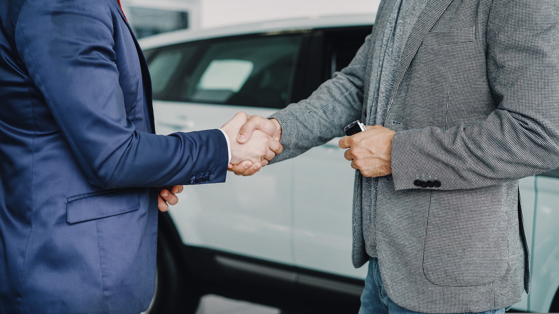 Male worker of car showroom is giving car keys to buyer young man and shaking hands with him standing beside luxurious new car. Selling and buying vehicles concept.